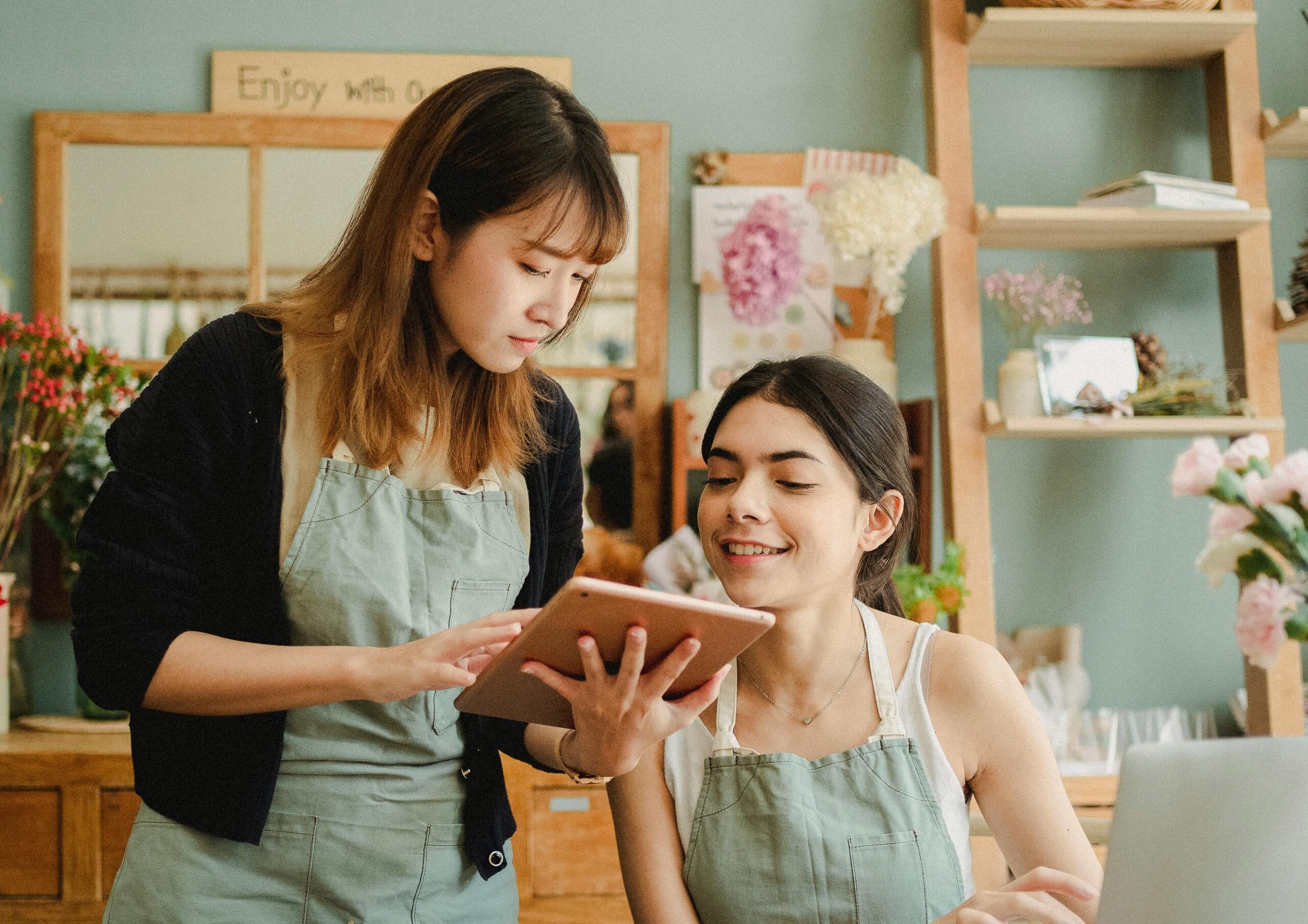 Concentrated multiethnic female florists wearing aprons at table with laptop and tablet checking orders online while working in flower store